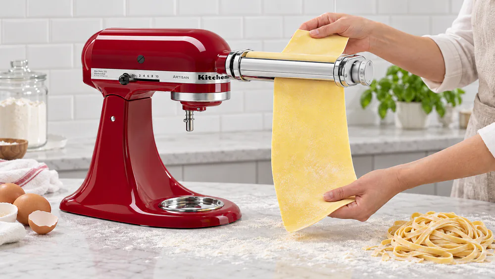 A sheet of fresh egg pasta passing through the KitchenAid pasta roller attached to a stand mixer, with a floured counter and a nest of cut fettuccine nearby