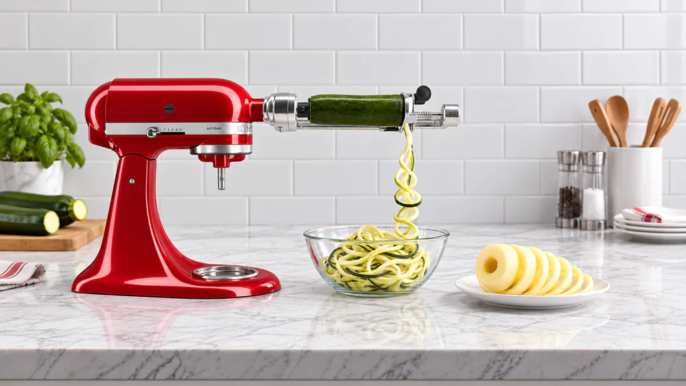 The KitchenAid 5-blade spiralizer attachment on a stand mixer turning a zucchini into noodles, with a peeled and cored apple spiral on a plate beside it