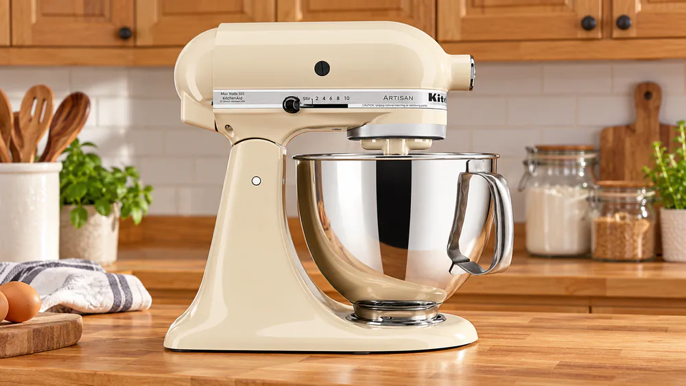 A warm creamy beige glossy Almond Cream KitchenAid Artisan stand mixer on a butcher-block counter with honey-toned wood cabinets behind it