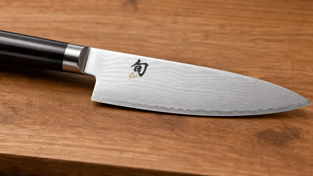 A close-up of a Shun Classic chef's knife showing the rippling Damascus cladding pattern along a VG-MAX core edge on a wooden cutting board.