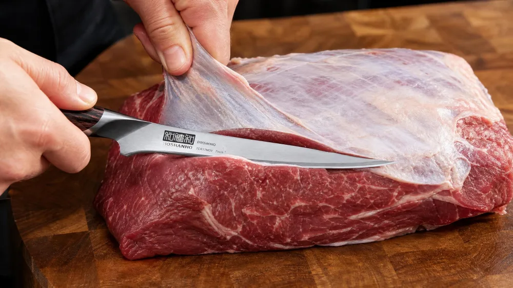 A HOSHANHO 7-inch fillet knife trimming silver skin from a large beef roast on a butcher block.