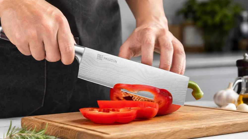 A PAUDIN 7-inch nakiri knife push-cutting a red bell pepper on a cutting board, the flat blade profile visible.