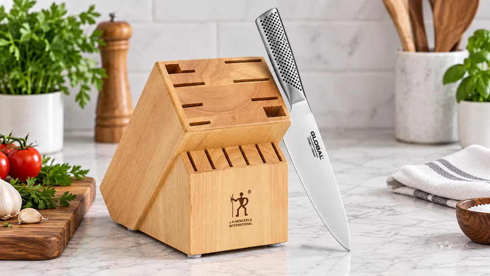 A Global chef's knife with its dimpled, sand-filled stainless handle leaning against the 11-slot stainless block.