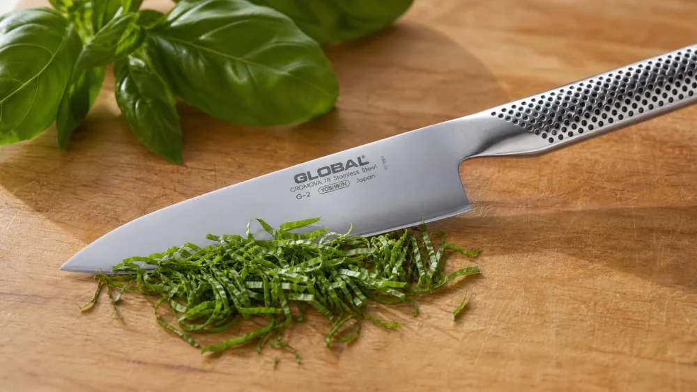 A Global G-2 chef's knife producing a fine chiffonade of basil, the dimpled one-piece steel handle and silver blade catching the light.