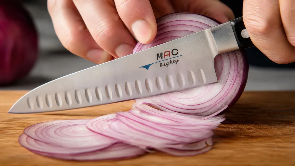 A Mac MTH-80 chef's knife slicing a red onion into paper-thin translucent slices on a wooden cutting board, the dimpled Granton edge visible along the blade.