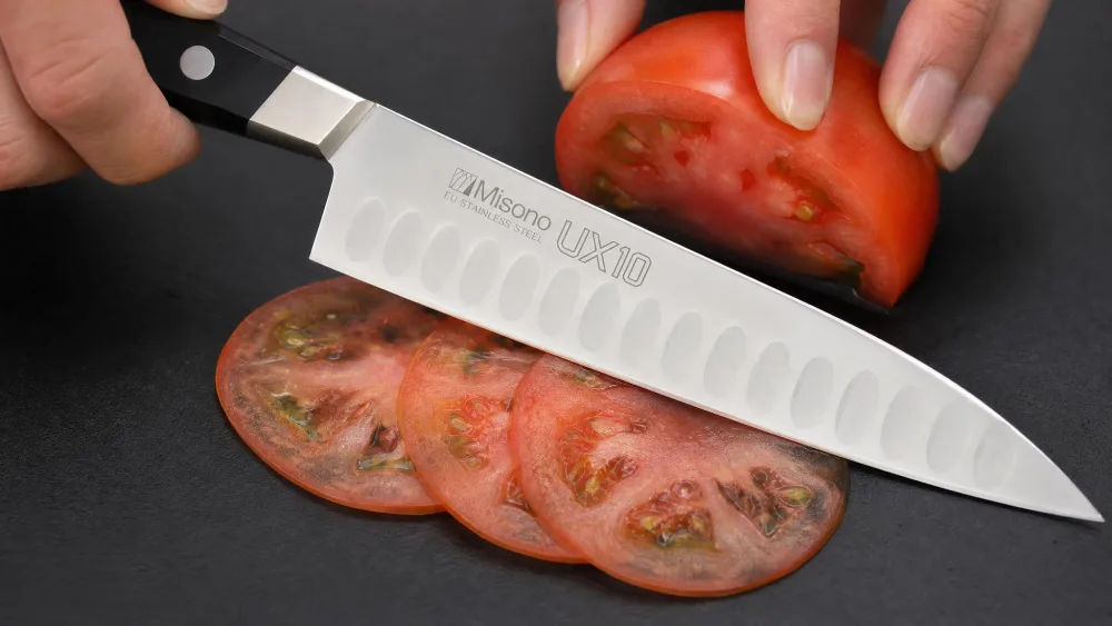 A Misono UX10 chef's knife cutting paper-thin translucent slices of tomato on a dark cutting board, the silver-nickel bolster and slim blade visible.