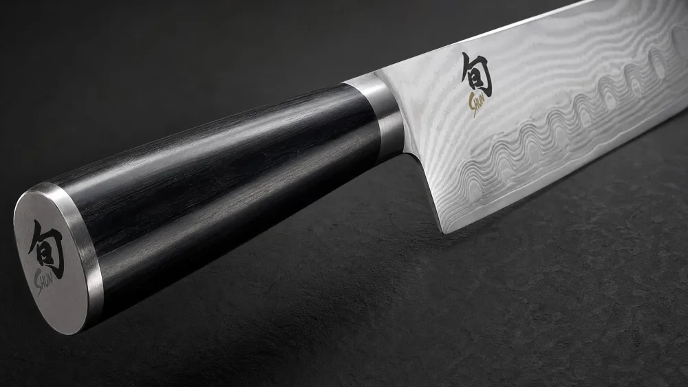 Close-up of a Shun Classic chef's knife showing the wavy 68-layer Damascus pattern along the blade, with the D-shaped Pakkawood handle in the foreground.