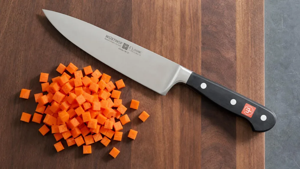 A Wüsthof Classic chef's knife on a cutting board beside a neat pile of evenly diced carrot cubes, the triple-riveted full tang and bolster visible.