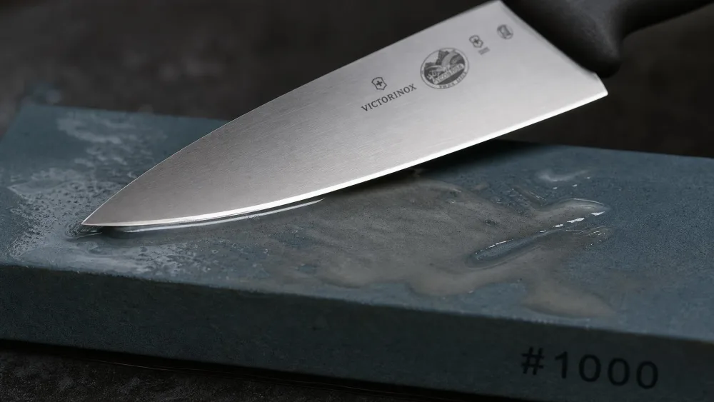 A chef's knife edge resting at an angle on a wet #1000 grit whetstone, the bevel catching the light and a slurry visible on the stone surface.