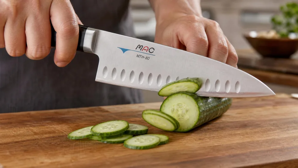 A MAC MTH-80 chef's knife mid-slice through a cucumber on a wooden board, thin rounds falling away from the dimpled hollow edge rather than sticking to the blade.
