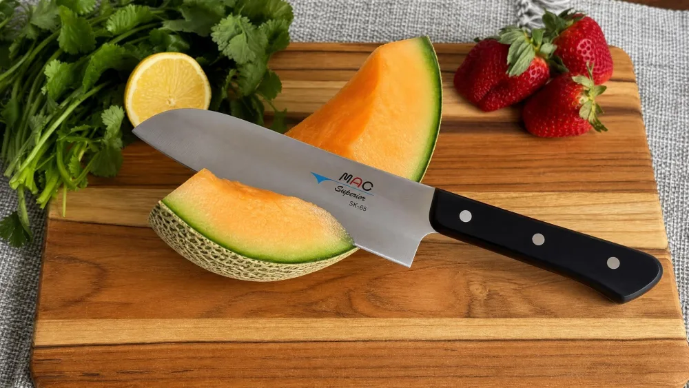 A Mac Superior Santoku SK-65 cutting through thick melon rind on a wooden board, the thin blade passing cleanly without wedging the halves apart.