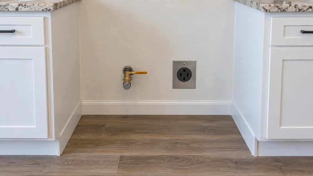 The back wall of a kitchen showing a gas shutoff valve and a 240V range outlet where a freestanding range will sit