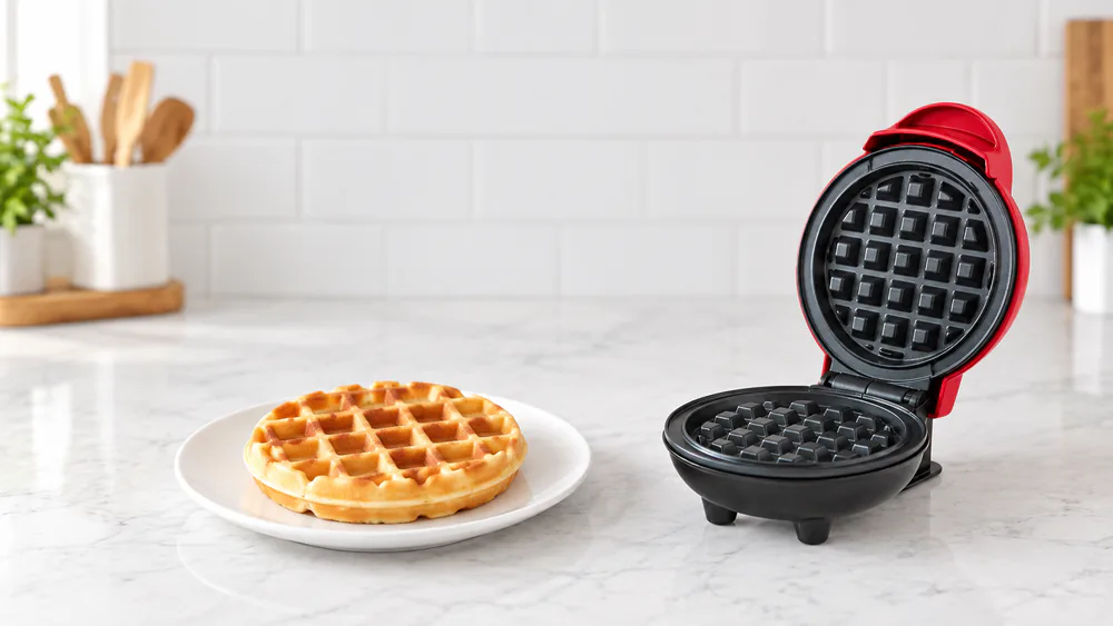 A golden 4-inch waffle on a white plate beside an open Dash Mini Waffle Maker