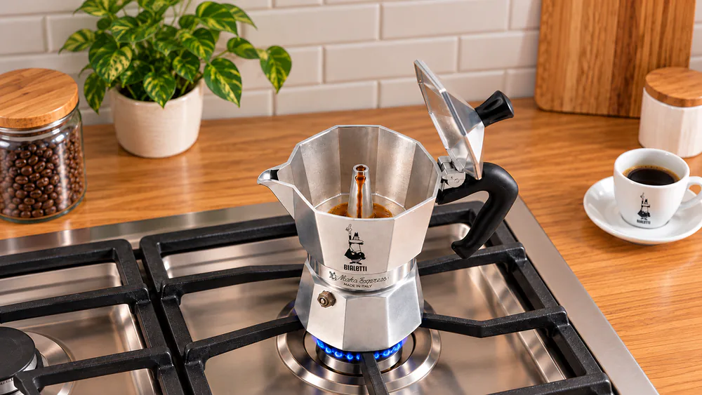 A Bialetti Moka Express 3-cup aluminum pot with its octagonal body sitting on a gas burner on low heat, lid open as coffee begins to flow into the collector