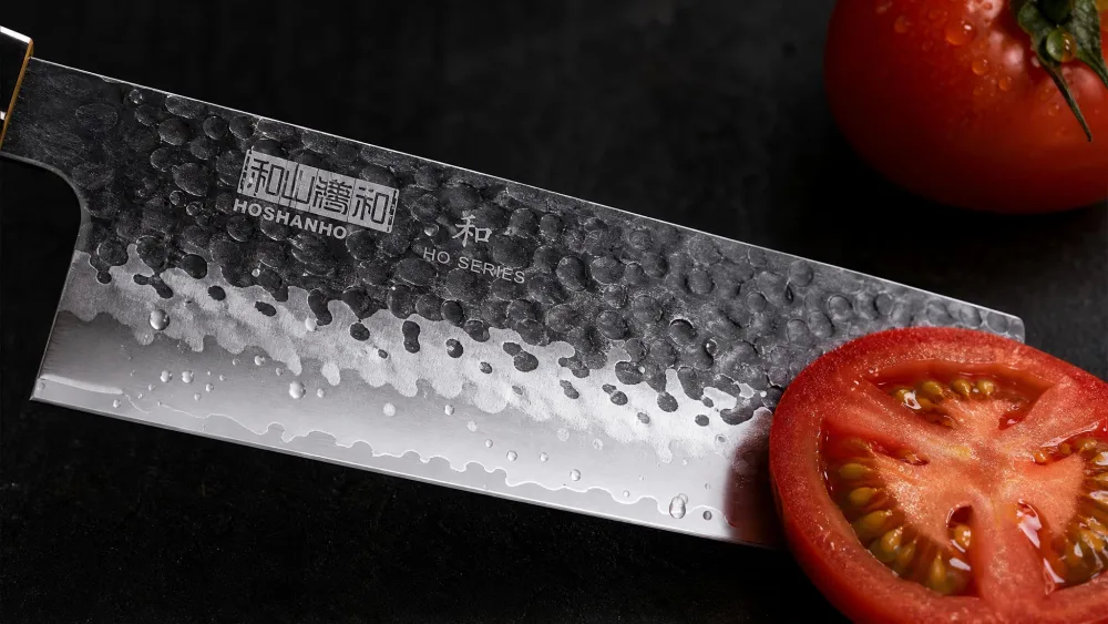 A close-up of the HOSHANHO nakiri blade showing the scallop-shaped hollow pits beside a tomato slice.