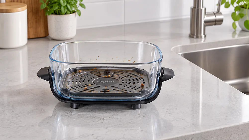 A used Ninja Crispi 4-qt glass container resting on its heat-safe feet on a quartz counter beside the sink, cooling before washing.