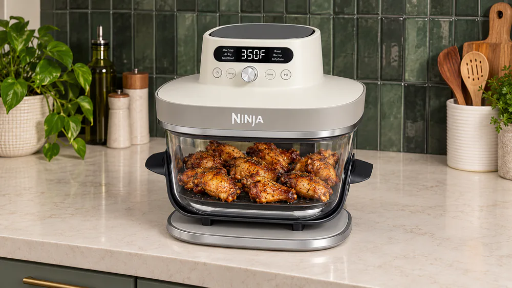 Browned chicken wings in a single layer inside the Ninja Crispi Pro 6-in-1's 6-qt glass container, seated in its stainless-steel base.