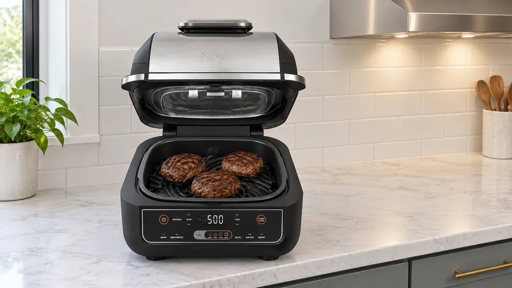 A Ninja EG201 indoor grill sitting on a kitchen counter beside a range hood vent with the lid open and burgers on the grate