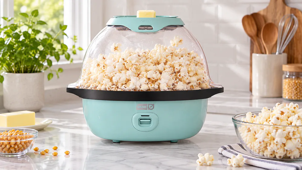 The Dash SmartStore Deluxe with its serving bowl lid filled with popcorn and the butter-warming tray on top, on a small apartment counter