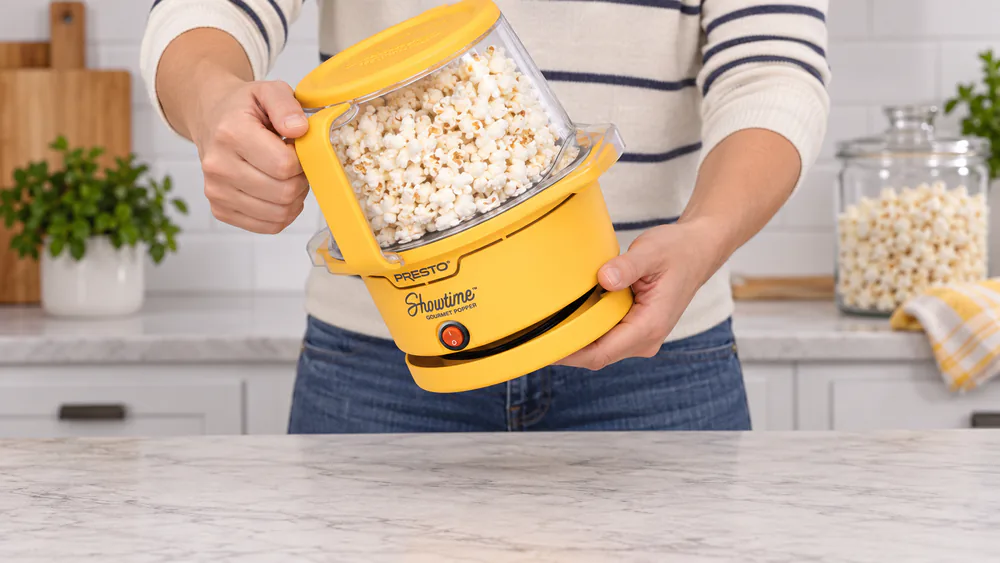 A hand flipping the Presto Showtime to finish a batch, popcorn tumbling under the transparent cover on a kitchen counter