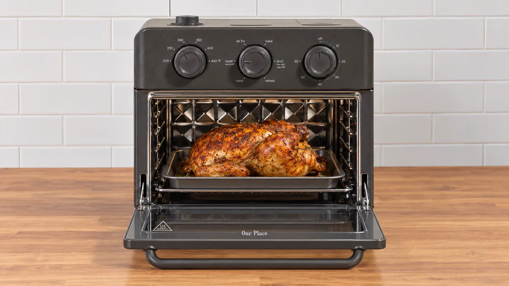 A roasted 4.5 lb chicken on the bake pan inside the standard Our Place Wonder Oven with the door open on a small kitchen counter