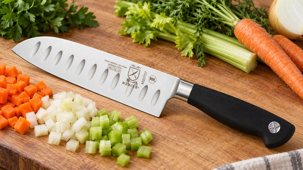 The Mercer Culinary Genesis 7-inch santoku knife with a black Santoprene handle and scalloped Granton blade beside a pile of diced vegetables.