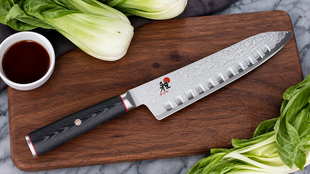 The Miyabi Kaizen 7-inch hollow edge santoku knife with a Damascus-pattern blade and D-shaped black linen Micarta handle.