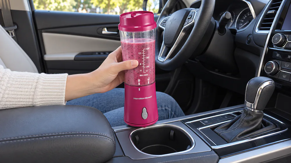 A Hamilton Beach personal blender jar with travel lid held beside a car cup holder to show its size.
