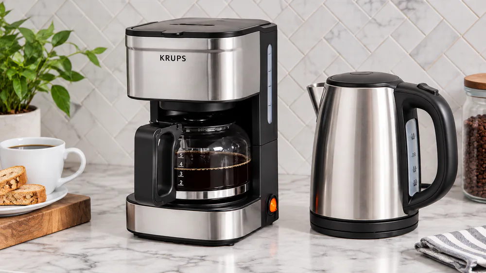 A compact KRUPS Simply Brew 5-cup drip coffee maker with its glass carafe filled, sitting on a narrow kitchen counter beside a kettle