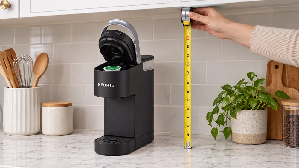 A tape measure held between a kitchen counter and the underside of an upper cabinet above a compact coffee maker with its lid open