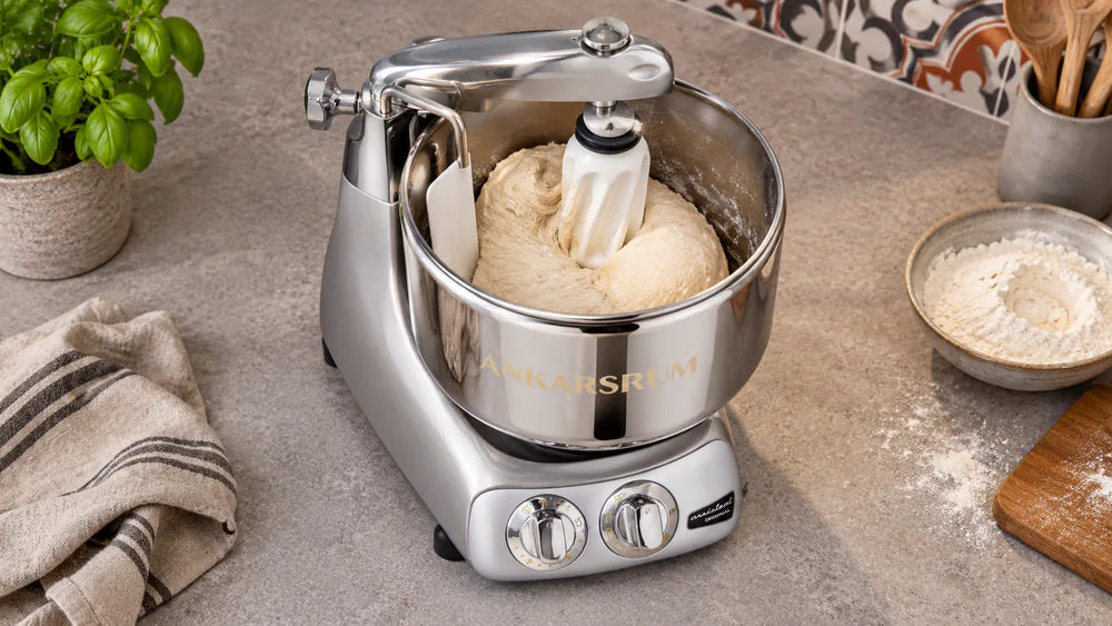An Ankarsrum Original mixer with its stainless bowl spinning and the dough roller and scraper working a ball of bread dough