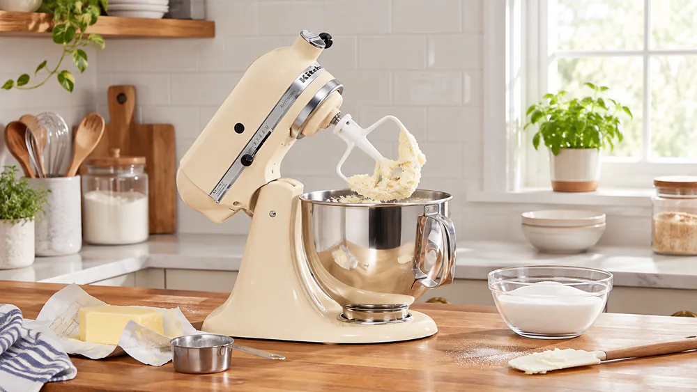 A KitchenAid Artisan tilt-head mixer on a kitchen counter with the head tilted back and a bowl of creamed butter and sugar inside