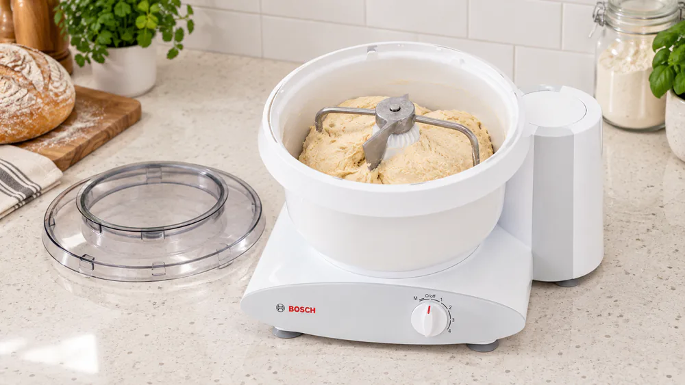 Bosch Universal Plus with a stiff ball of bread dough wrapped around the center post of its plastic bowl, suction-cup feet on the counter
