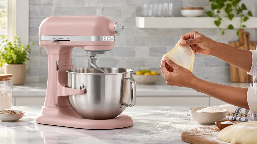 Baker stretching a piece of dough into a thin windowpane beside a KitchenAid 7-Qt bowl-lift mixer with its stainless bowl lowered