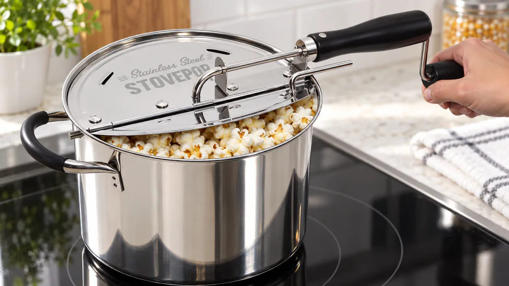 A Victorio StovePop stainless popper on a gas stove, crank handle raised to show the gearless flex-drive cable.