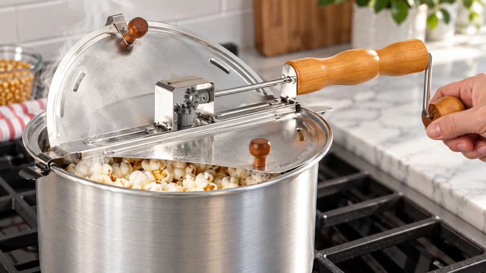 An Original Whirley Pop crank popper on a glass-top stove, lid off, showing the patented stirring mechanism and four steam vents.
