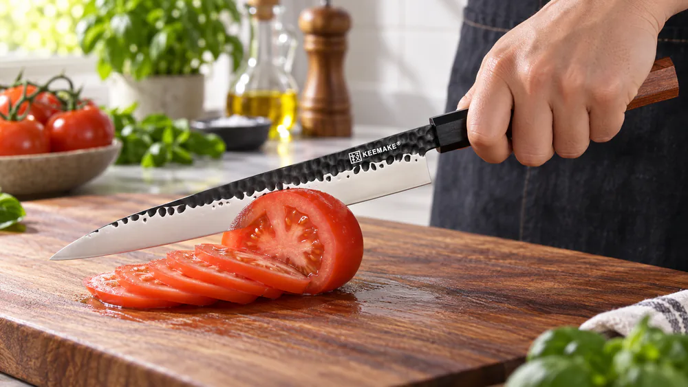 A KEEMMAKE yanagiba slicing a ripe tomato cleanly on a wooden board, hydrophobic black coating on the blade.