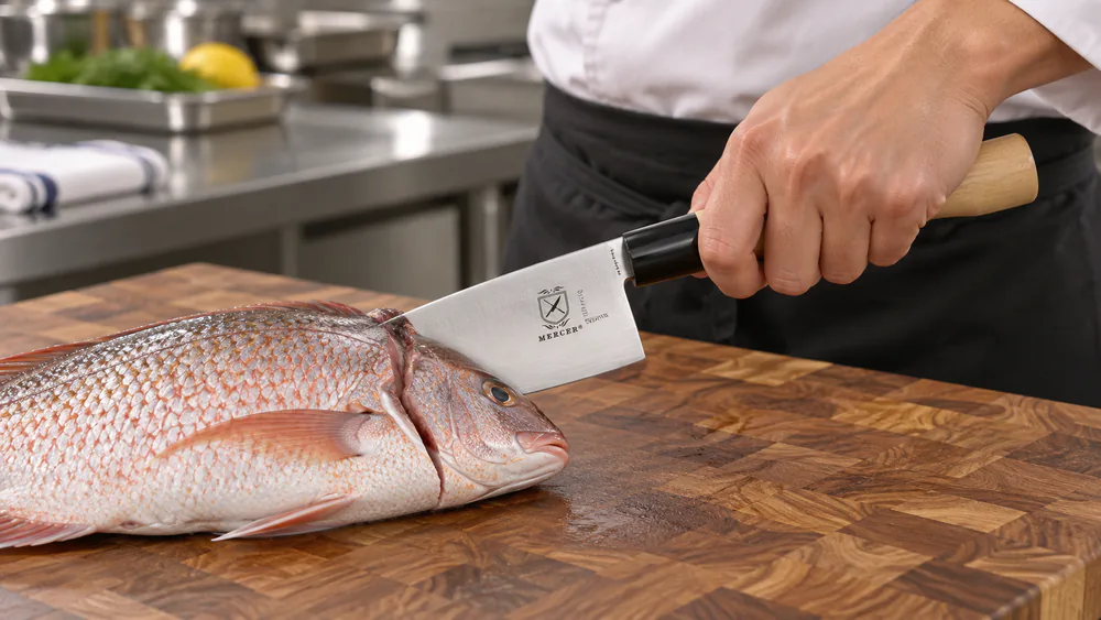 A Mercer Culinary deba knife breaking down a whole fish on a commercial kitchen cutting board.