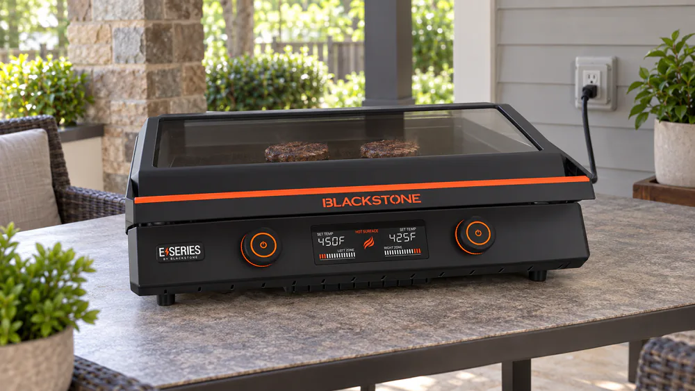A Blackstone E-Series 22 inch electric griddle with the hood on, plugged in and cooking on a patio table