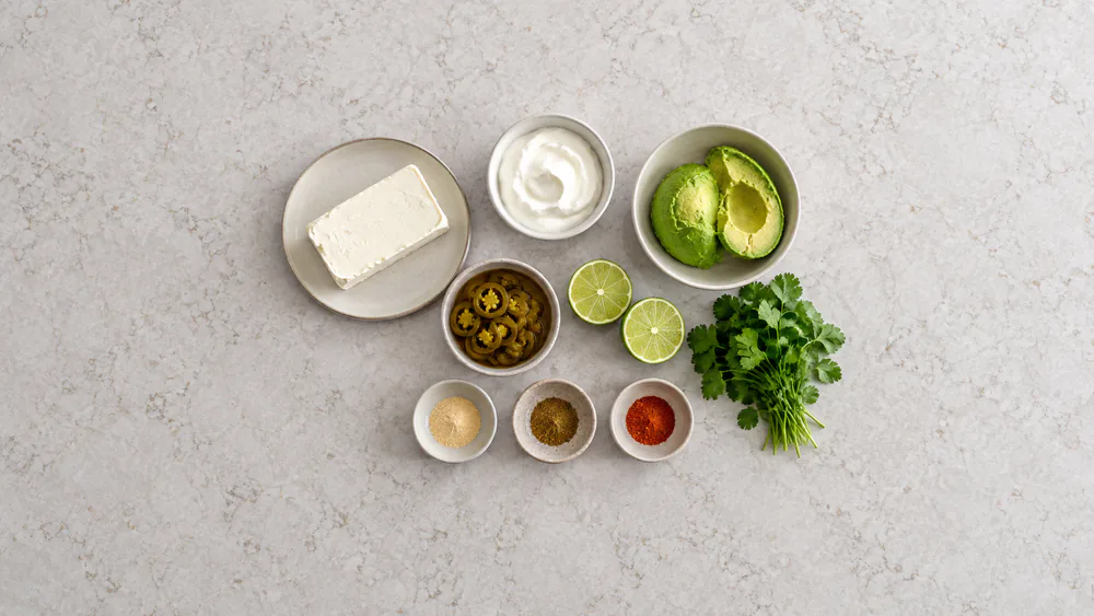 All ingredients for the Cardi B avocado jalapeño dip laid out flat including cream cheese, sour cream, avocado, pickled jalapeños, lime, and cilantro