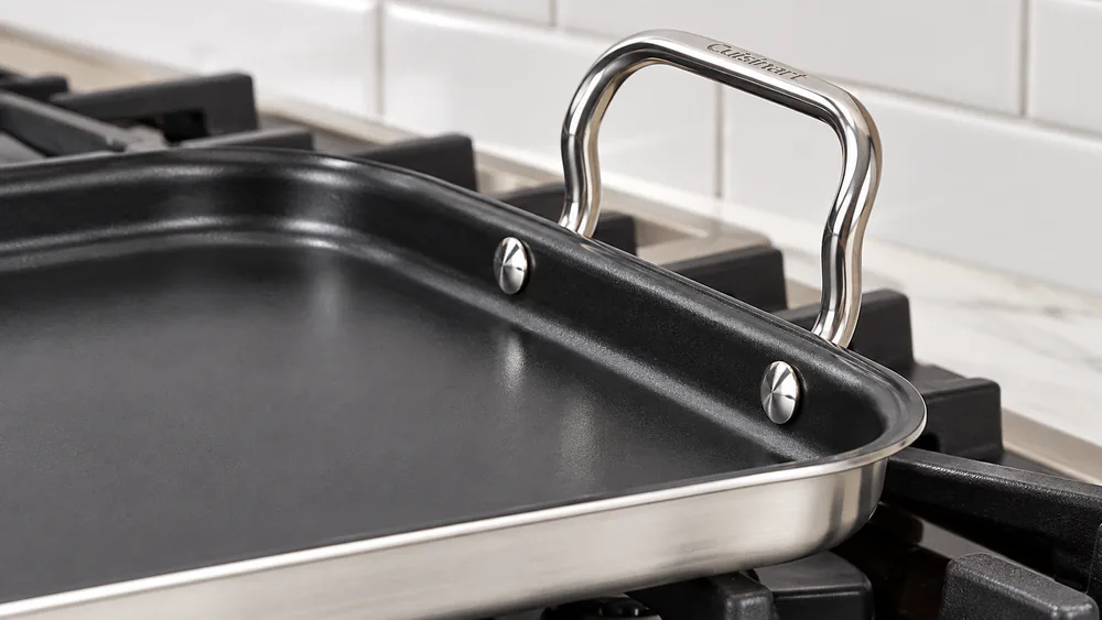 Close-up of the riveted stainless Cool Grip handle and drip-free pouring rim on the Cuisinart double burner griddle sitting on a gas range