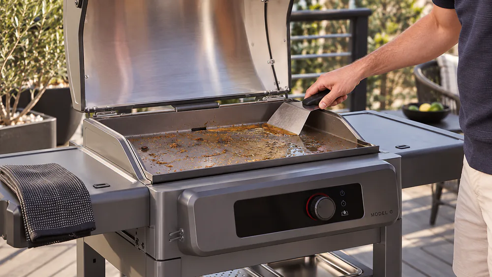 A metal scraper pushing grease toward the rear grease tray of the Current Model G Electric Griddle after cooking