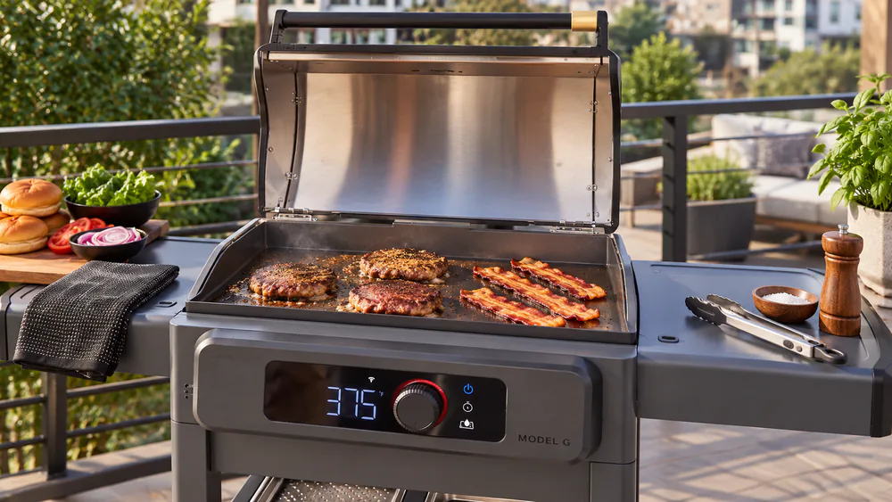 Smash burgers and strips of bacon cooking side by side on the seasoned steel surface of the Current Model G Electric Griddle