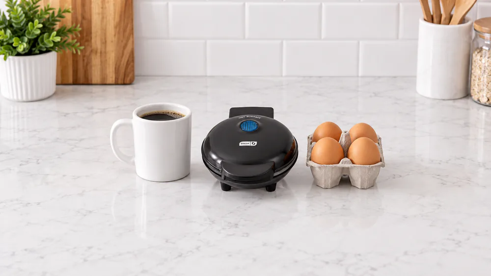 The black Dash Mini Maker round griddle closed on a small kitchen counter next to a mug and a carton of eggs