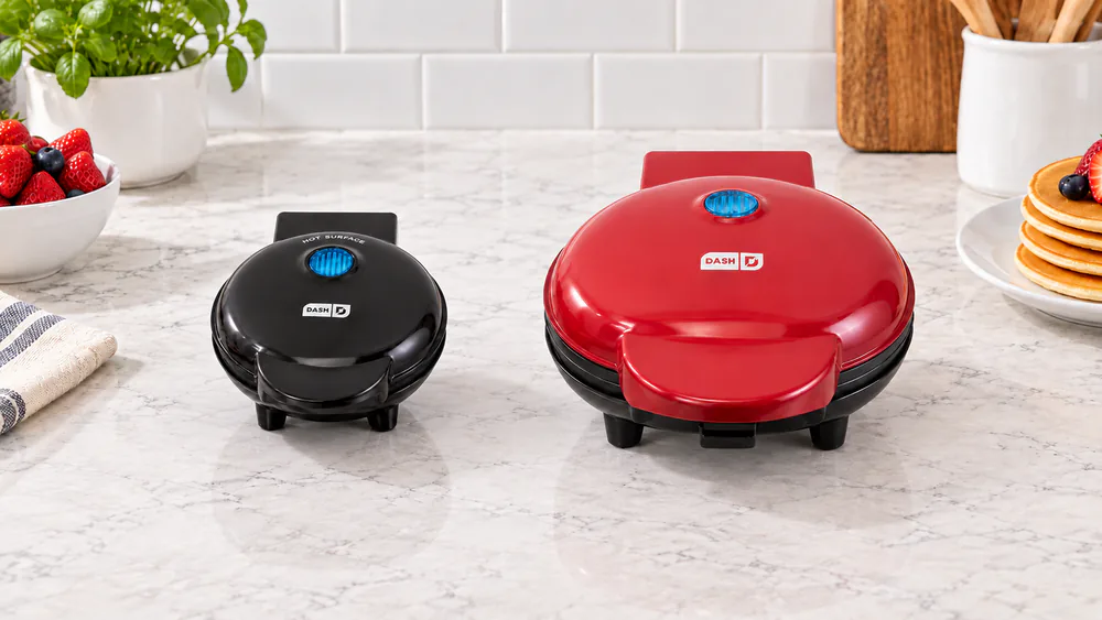 The 4-inch Dash Mini Maker sitting beside the larger 8-inch Dash Express griddle on a kitchen counter for size comparison