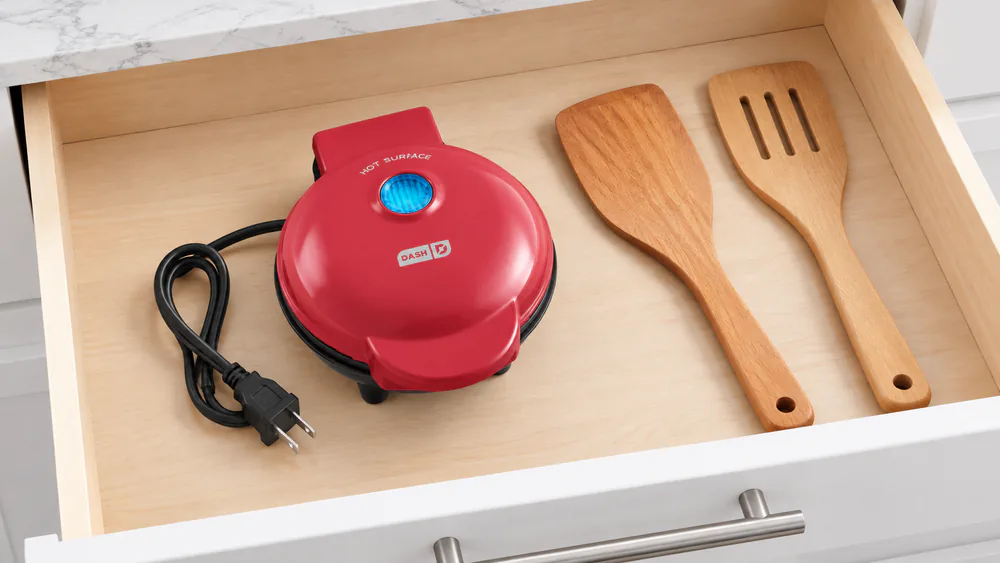 A compact Dash Mini waffle maker tucked into a kitchen drawer beside spatulas, with its short cord wrapped beside it
