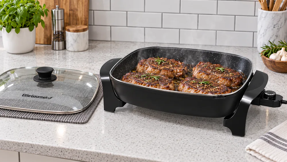 An Elite Gourmet EG-6203 electric skillet with several steaks searing across the center of the deep 16-inch nonstick pan, steam rising, glass lid resting to the side.