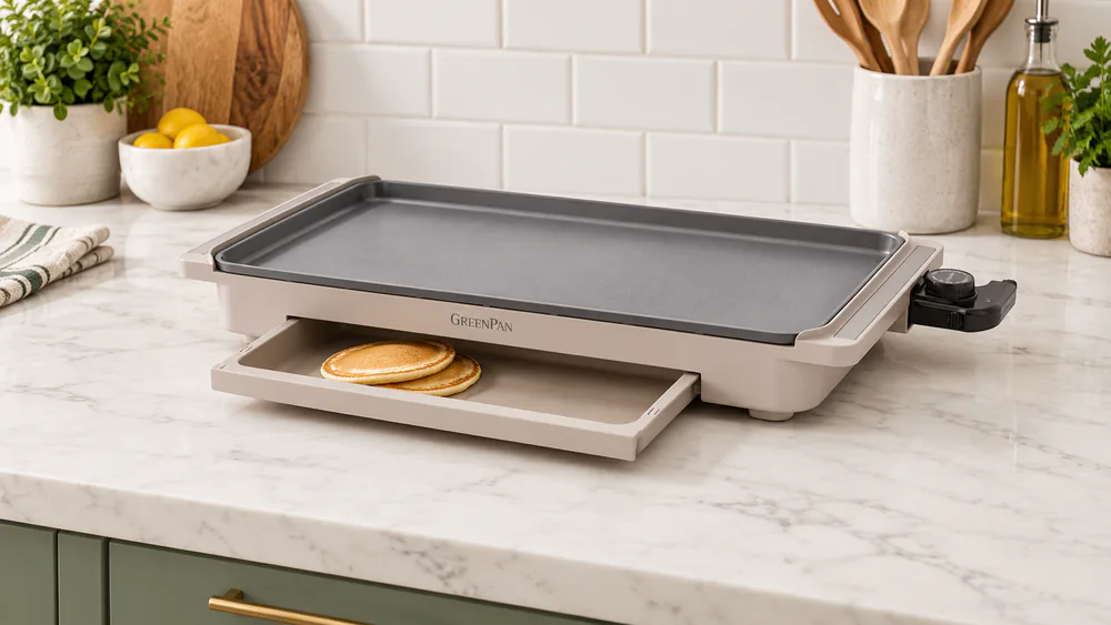 The Clay GreenPan Advanced electric griddle on a kitchen counter with the warming drawer slightly open and a stack of pancakes inside