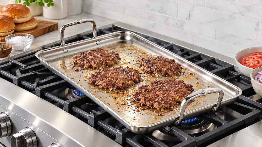 A row of smash burgers with browned crusts cooking on the brushed stainless surface of the Hestan Provisions griddle over two gas burners