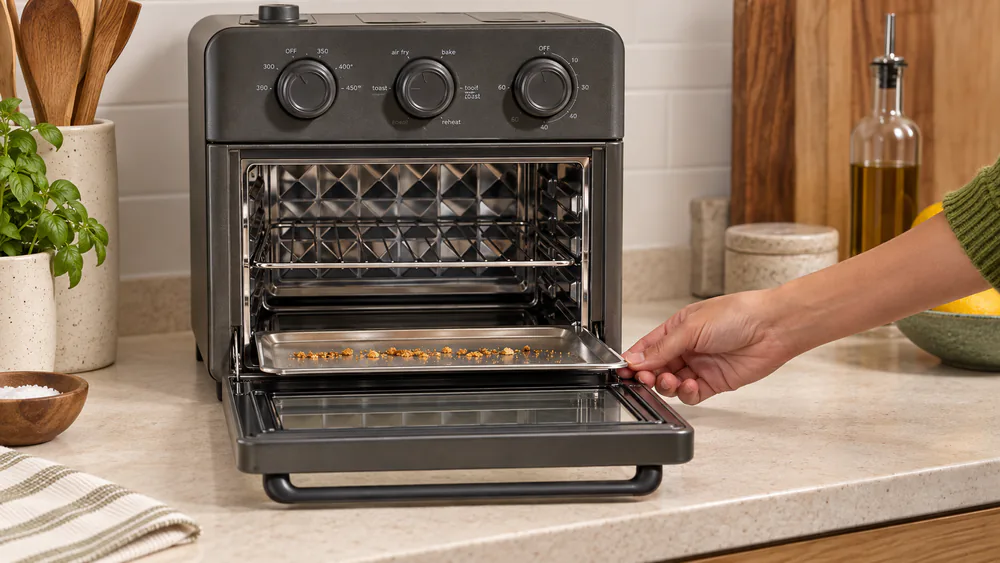 The stainless steel crumb tray sliding out from beneath the bottom heating element of an Our Place Wonder Oven, crumbs gathered at its edge.